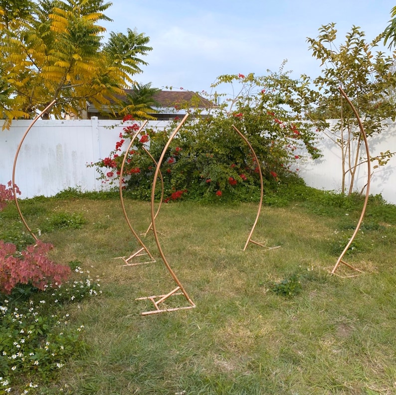 May include: Five copper-colored metal arch stands are arranged on a grassy lawn. Each stand has a curved top and a base with a geometric design. The stands are outdoors, with a white fence and greenery in the background.