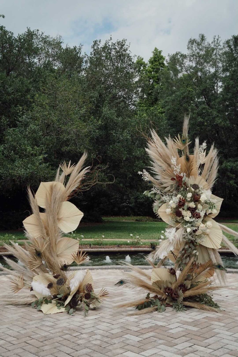 May include: Two decorative floral arrangements featuring dried palm leaves, pampas grass, and various dried flowers. The arrangements are in shades of beige, brown, and white, set against a backdrop of greenery and a water feature.