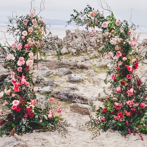 May include: Two floral arches, adorned with pink and red roses, stand on a rocky beach. The arches are decorated with greenery and branches, creating a natural and romantic setting. The background features a body of water and a cloudy sky.