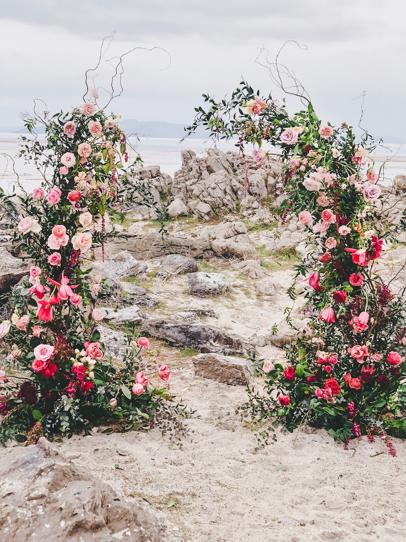 May include: Two floral arches, adorned with pink and red roses, stand on a rocky beach. The arches are decorated with greenery and branches, creating a natural and romantic setting. The background features a body of water and a cloudy sky.