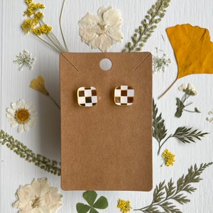 Puede incluir: Un par de pendientes cuadrados con borde dorado y un patrón de cuadros blancos y negros. Los pendientes se exhiben en una tarjeta marrón, rodeados de flores y hojas secas sobre una superficie blanca.