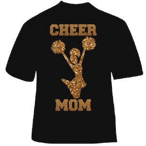 May include: Black t-shirt with gold glitter design. The design features the words "CHEER MOM" above and below a cheerleader silhouette holding pom-poms. The text and cheerleader are in a gold glitter finish.