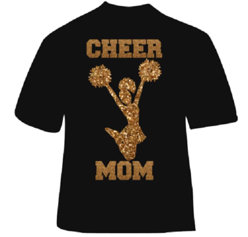 May include: Black t-shirt with gold glitter design. The design features the words "CHEER MOM" above and below a cheerleader silhouette holding pom-poms. The text and cheerleader are in a gold glitter finish.