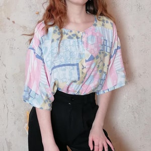 Könnte beinhalten: Eine kurzärmelige, geknöpfte Bluse mit einem geometrischen Muster in Rosa-, Blau- und Gelbtönen. Die Bluse hat eine lockere Passform und einen runden Ausschnitt.