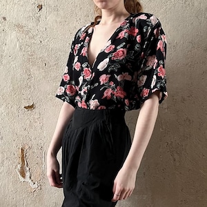 Op de afbeelding: Een zwart bloemetjesblouse met een diepe V-hals en korte mouwen. Het blouse wordt gecombineerd met een zwarte broek met hoge taille.