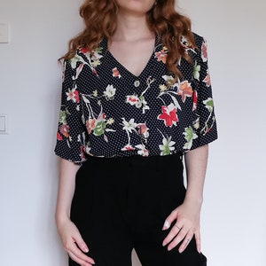 Könnte beinhalten: Eine schwarz-weiße Polka-Dot-Bluse mit einem Blumendruck in Rot-, Gelb- und Grüntönen. Die Bluse hat einen V-Ausschnitt und kurze Ärmel.