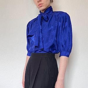 Könnte beinhalten: Eine Vintage-Bluse in Kobaltblau mit hohem Kragen und Schleife. Die Bluse hat ein dezentes Rosenmuster und kurze, gepuffte Ärmel. Kombiniert mit einem hoch taillierten schwarzen Rock, wodurch ein klassischer, eleganter Look entsteht.