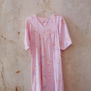 Könnte beinhalten: Ein rosa Nachthemd mit Blumenmuster und kurzen Ärmeln, das an einem weißen Kleiderbügel an einer strukturierten Wand hängt.