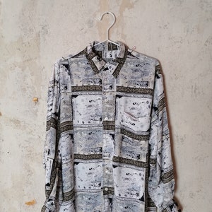 Puede incluir: Una camisa de manga larga con botones, fondo blanco y diseño estampado en negro, gris y verde. El estampado presenta un diseño repetitivo de formas geométricas y figuras abstractas.