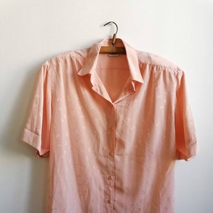 Könnte beinhalten: Ein pfirsichfarbenes Hemd mit Knöpfen, kurzen Ärmeln und einem Blumenmuster. Das Hemd hängt an einem Holzbügel.