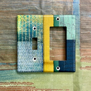 May include: A light switch cover with a colourful abstract design. The cover is made of plastic and has a rectangular shape. The design features shades of blue, green, yellow, and white.