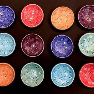 Puede incluir: Doce cuencos pequeños de cerámica, cada uno con una letra diferente del alfabeto. Los cuencos están dispuestos en una cuadrícula y son de varios colores, incluidos azul, verde, morado, rojo y naranja.