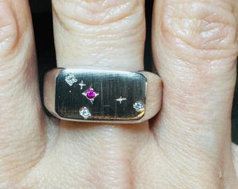 May include: A silver signet ring with a rectangular face. The face features a design of a pink gemstone and several small clear gemstones. The ring is worn on a finger.
