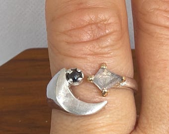 May include: A silver-toned open ring featuring a crescent moon shape on one end set with a small dark stone and a diamond-shaped clear stone set in gold-toned prongs on the other end. The band is smooth and wraps around the finger with an open gap.