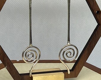 May include: Silver-toned dangle earrings with a unique spiral design. The earrings have a long, slender bar that ends in a decorative spiral. The earrings are displayed on a wooden stand with a gold-colored base.