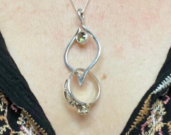 May include: A silver necklace with a pendant featuring a swirling design and a green gemstone. The pendant is attached to a ring with a smaller gemstone.