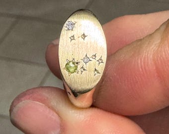 May include: Gold-toned oval signet ring with a celestial design. The ring features a small, round, light green gemstone and several diamond-like accents. The ring is held between fingers.