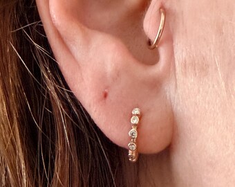 May include: A gold hoop earring featuring a row of five small, clear, round-cut stones set along the front, positioned in the earlobe, accompanied by a plain, thin gold hoop earring worn in the tragus area of the ear.