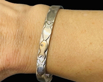 May include: A silver bangle bracelet with a repeating pattern of fish. One fish is highlighted in gold with a small diamond.