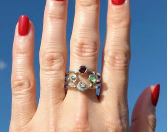 May include: A multi-band silver ring featuring a dark red circular gemstone at the top, two smaller light green circular gemstones on the sides, and a small crescent moon shape with a tiny embedded clear stone at the bottom of the ring design.