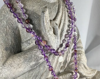 May include: A long beaded necklace featuring translucent purple and clear quartz beads with reddish inclusions, strung on a vibrant purple cord. The necklace includes small silver-toned spacers, a circular charm, and a light-colored tassel at the end.