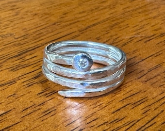 May include: A silver-toned ring with a spiral design. The ring features a small, round, clear stone at the top. The band is made of multiple hammered layers. The ring is displayed on a wooden surface.