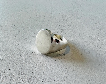 May include: A silver signet ring with an oval face. The ring is made of polished metal and sits on a textured, light gray surface. The ring's design is simple and classic.