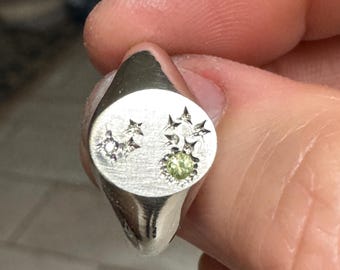 May include: A silver signet ring with a brushed finish. The ring features a design of stars and a small, round, light green gemstone. The ring is held in a person's hand.
