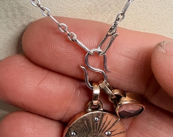 May include: A silver chain necklace featuring an S-hook clasp that holds two pendants. One pendant is a round, gold-toned disc with engraved sunburst lines and small embedded stones. The second pendant is a small, teardrop-shaped, purple-toned stone.