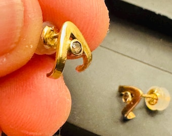 May include: A pair of gold stud earrings shaped like the letter A, each featuring a small, clear, round-cut gemstone set into the center of the letter. The earrings include translucent plastic earring backs for secure attachment to the earlobe.