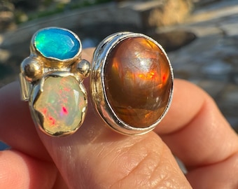 May include: Two gemstone rings. One ring features a large, oval, fiery orange stone set in a silver band. The other ring has a gold band and two stones: a blue-green oval and a white opal with flashes of color.