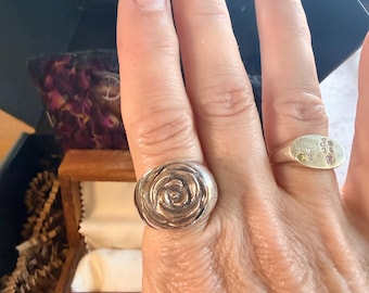 May include: A silver ring featuring a sculpted rose design on the index finger and a silver oval signet ring with small embedded stones on the ring finger. Both rings have a polished metallic finish and are worn on the same hand.