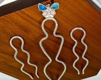 May include: Three silver hair pins with a wavy, elongated silhouette. The center pin features a butterfly-shaped ornament at the top, inlaid with two blue iridescent stones and two smaller, pale pink stones set in a polished silver frame.