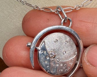 May include: A silver pendant necklace featuring a circular frame that holds a rotating disc. The disc has a textured surface with a crescent moon shape, small circular indentations, and three small embedded clear stones set in the metal.