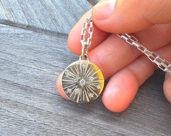 May include: A circular silver pendant featuring a sunburst pattern with small embedded clear stones, suspended from a rectangular link chain. The metal surface has a polished finish with radiating lines extending from the center point.