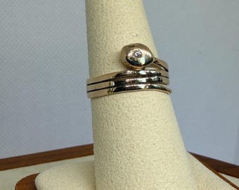 May include: A gold-colored ring with a small diamond set in a rounded top. The ring has a coiled design with three bands and is displayed on a cream-colored ring holder.