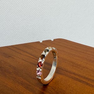 May include: A silver ring featuring a line of colourful gemstones. The gems are in shades of red, pink, brown, green, and yellow. The ring is displayed on a wooden surface.