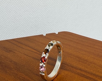 May include: A silver ring with a row of colorful gemstones. The gemstones are in shades of red, pink, brown, green, and yellow. The ring is set on a wooden surface.