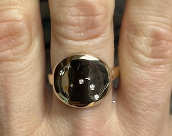 May include: A gold ring with a round, slightly domed top set with small diamonds. The ring is worn on a finger.