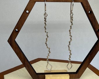 May include: A pair of silver-toned earrings with a twisted wire design. Each earring has a long, spiraling body and a small, diamond-shaped charm at the bottom. The earrings are displayed on a wooden stand.