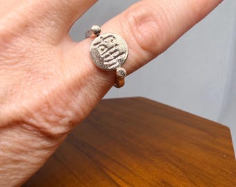 May include: A silver-toned ring featuring a circular centerpiece with a textured surface and embossed lettering. The band is thin and includes two small spherical accents where it connects to the central circular disc, creating a distinct design.