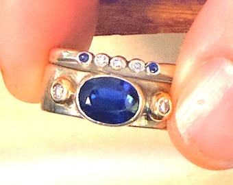 May include: A two-piece ring set featuring a wide band with a large oval blue sapphire center stone flanked by two small round diamonds in gold settings. The second band is a thinner silver-toned ring with a row of alternating blue and white stones.