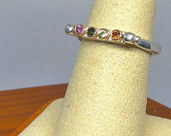 May include: A silver band ring featuring four small, round, bezel-set gemstones in a row. The stones are colored pink, dark green, light green, and orange. The ring has a polished metallic finish and a smooth, rounded band design.