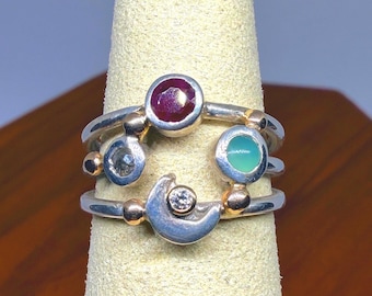 May include: A multi-band ring featuring a silver and gold construction with a round faceted deep red stone, a round faceted dark grey stone, a round smooth light green stone, and a crescent moon shape accented with a small clear round stone.