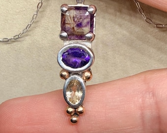 May include: A vertical pendant necklace featuring a square rutilated quartz, an oval amethyst, and a smaller oval citrine set in silver. The design includes small gold-toned metal spheres accenting the lower stones, suspended on a silver chain.