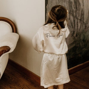 Op de afbeelding: Een jong meisje draagt een witte satijnen badjas met de tekst "Flower Girl" erop gedrukt. Ze staat voor een donkergrijze muur.