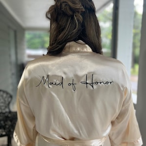 Op de afbeelding: Een cr&egrave;mekleurige satijnen badjas met de tekst "Maid of Honor" geborduurd in het zwart op de rug.