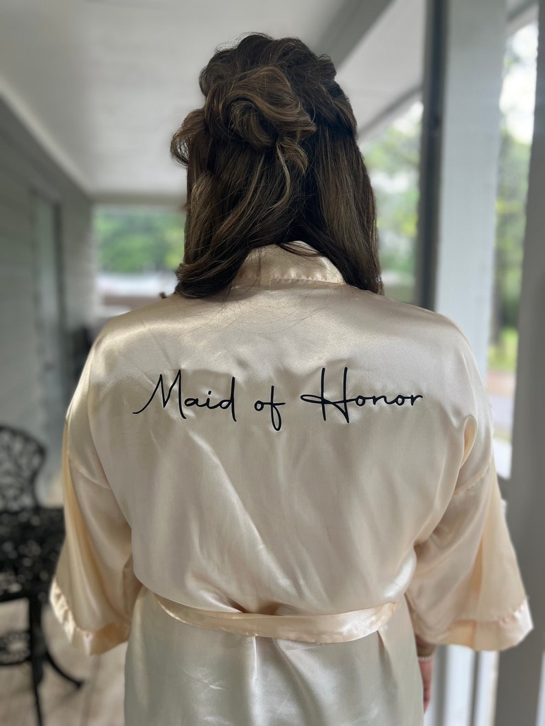 Op de afbeelding: Een cr&egrave;mekleurige satijnen badjas met de tekst "Maid of Honor" geborduurd in het zwart op de rug.