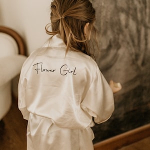 Op de afbeelding: Een witte satijnen badjas met de tekst "Flower Girl" geborduurd op de rug.