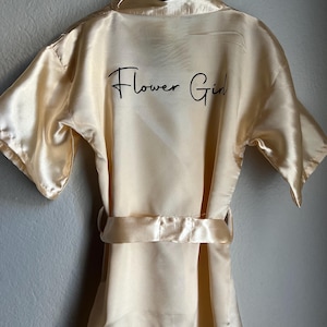 Op de afbeelding: Een cr&egrave;mekleurige satijnen badjas met korte mouwen en een ceintuur. De badjas heeft de woorden "Flower Girl" in het zwart geborduurd op de achterkant.